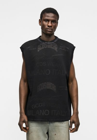 Νέος άνδρας με σκούρο δέρμα, που φοράει φαρδιά μαύρη αμάνικη μπλούζα με ξεθωριασμένο κείμενο και ανοιχτό γκρι τζιν, μπροστά σε απλό ανοιχτόχρωμο φόντο.