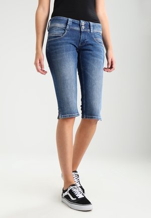 Kvinde iført mid-lange blå denimshorts med dobbelt-knaplukning og sort sneakers med hvide snørebånd.