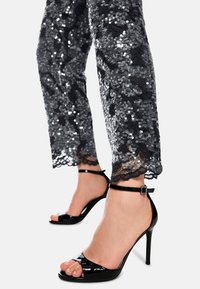 Legs wearing black sequined pants and black patent leather high-heeled sandals with ankle straps against a white background.