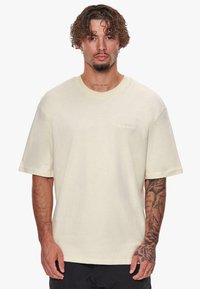 T-shirt à manches courtes de couleur crème clair, fabriqué en tissu doux, avec un col rond côtelé et un logo de la marque subtilement brodé sur la poitrine.