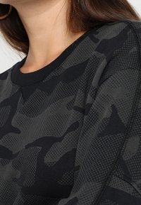 Mustat camouflage-kuvioiset collegepaidat, jotka on valmistettu tekstuurilla varustetusta kankaasta, jossa on ribbireunus kauluksessa ja näkyviä ompelutietoja hihoissa.