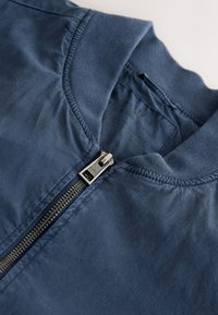 Blouson bomber bleu marine avec col côtelé et fermeture éclair argentée partiellement fermée, montrant le texte imprimé de l’étiquette intérieure et le logo de la marque sur la tirette.