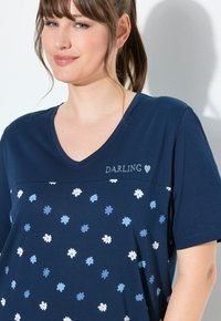 T-shirt a V blu navy con un motivo floreale in azzurro chiaro e bianco. Presenta la parola "DARLING" ricamata in bianco sopra il motivo.