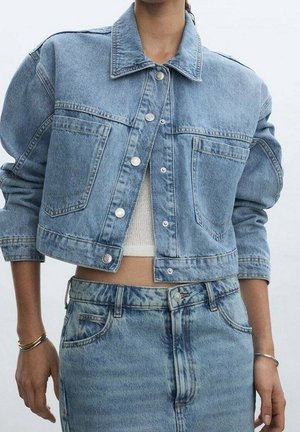 Femme portant une veste en jean courte bleu clair sur un top court blanc, associée à un jean bleu taille haute et des bracelets argentés aux deux poignets.