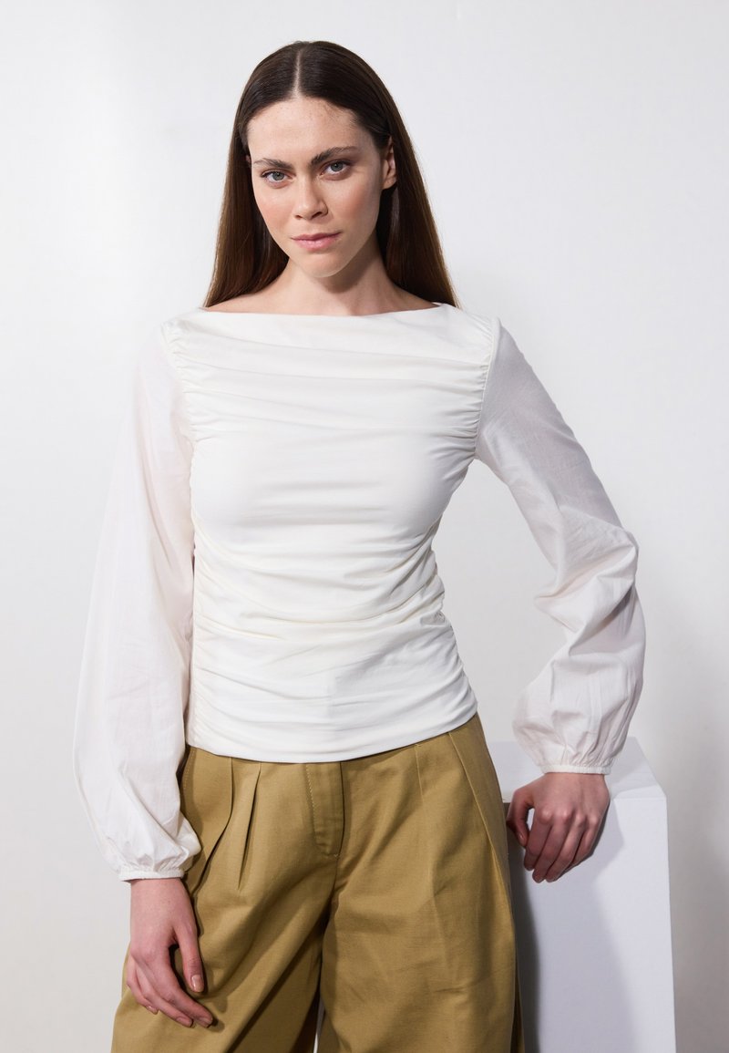 Hvit topp med ruchert byste og gjennomsiktige lange ermer, parret med khaki vide bukser. Glatt tekstur; minimalistisk design.