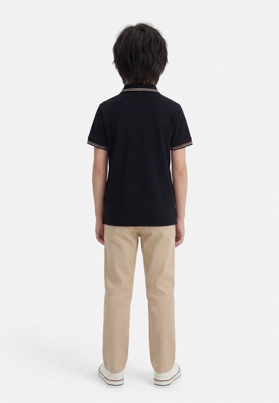 Παιδί που στέκεται με την πλάτη γυρισμένη, φορώντας μαύρο κοντομάνικο πόλο, μπεζ παντελόνι και λευκά αθλητικά παπούτσια, σε απλό λευκό φόντο.