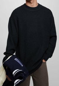 Pull en maille foncée avec une texture côtelée et un col rond, porté par une personne tenant un sweat-shirt bleu marine plié et une veste beige.