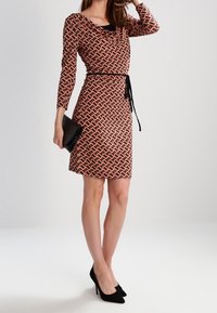 Czerwona i czarna sukienka w wzory z długimi rękawami, taliowana z paskiem. Teksturowana tkanina i dopasowany krój. Czarna kopertówka i szpilki.