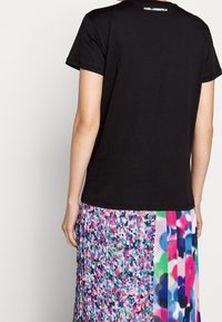 Camiseta de algodón negra de mangas cortas y cuello redondo, con un logotipo blanco en la parte posterior. Combinada con una falda de colores multicolores y con estampado.