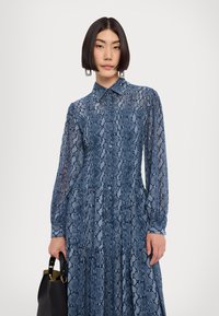 Rochie albastră cu imprimeu de șarpe, cu mâneci lungi, închidere cu nasturi și fustă plisată. Material ușor și model texturat. Ține în mână o geantă neagră.