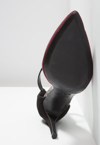 Chaussure à talons aiguilles noirs et bordeaux à bout pointu vue de la semelle avec un talon stiletto fin, posée sur une surface blanche devant un mur blanc.