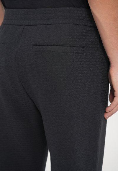 Zbliżenie na czarne spodnie z subtelnym, powtarzającym się wzorem logo Emporio Armani oraz tylnymi kieszeniami z patkami, noszone przez osobę z ręką swobodnie opuszczoną po boku.