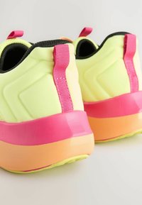 Neon žute sportske cipele s rozim naglaskom na peti i teksturiranim crnim obrubom. Potplat prelazi iz roze u narandžastu, pružajući efekt gradijenta.