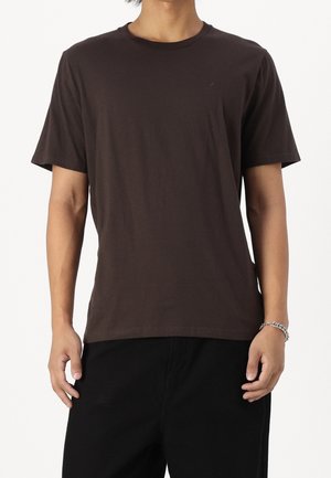 Persoon draagt een effen donkerbruine korte mouw T-shirt en zwarte broek, staat met ontspannen handen langs het lichaam tegen een witte achtergrond.