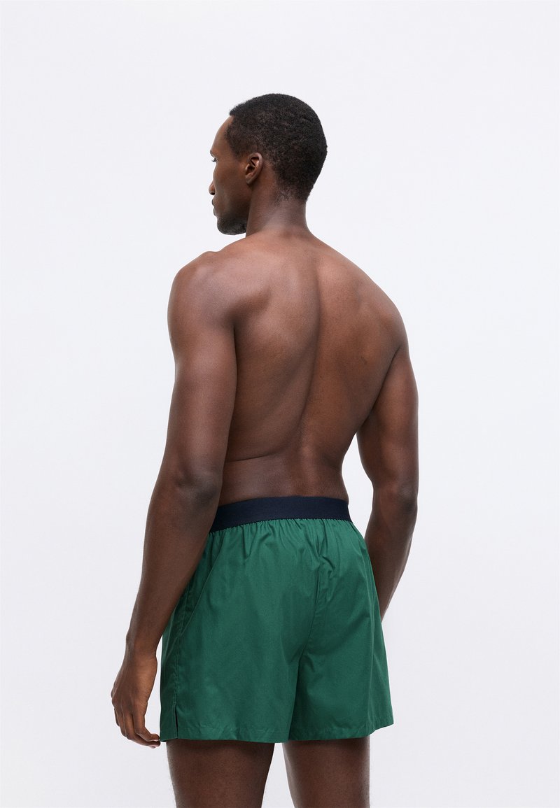 Green swim shorts with an elastic navy waistband. The fabric is smooth and lightweight. The design is loose-fitting with side seams.