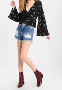 Una donna indossa una blusa nera a maniche lunghe con fiori, pantaloni corti in denim strappati e stivaletti alla caviglia di un rosso scuro lucido, posando su uno sfondo bianco.