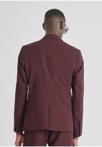 Giacca da suit borgogna con una texture liscia, spacchetto singolo sul retro e polsini abbottonati. Linee pulite e vestibilità su misura.