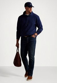 Marinblå hoodie med dragsko, mörkblå jeans i denim, brun läderväska, bruna sneakers, matchande mörk keps och klocka, allt i en avslappnad outfit.