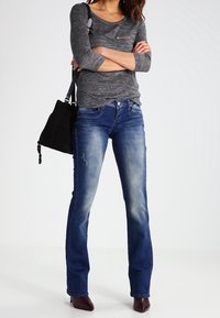 Maglione lungo a maniche grigio scuro con taschino sul petto, abbinato a jeans blu a campana con dettagli sbiaditi. Il modello tiene una borsa nera e indossa dei tacchi.