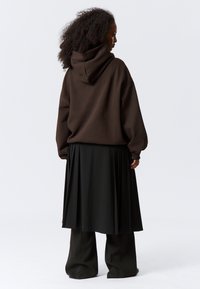 Sudadera marrón de talla grande con capucha, combinada con una larga falda negra plisada y pantalones anchos negros, mostrando texturas en capas.