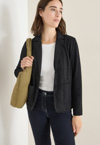 Sort tweedblazer med spidse lapler og lomme-detaljer, parret med en hvid top og mørke jeans. Modellen bærer en olivengrøn taske.