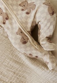 Pijama para bebé color crema con estampados de ositos marrones, tumbado sobre una manta beige texturizada, mostrando cremallera y pies cubiertos.