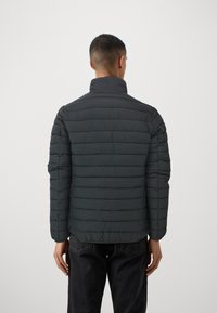 Puffjakke i mørkegrå med quiltet struktur, h�øj krave og justerbar pasform. Kombineret med sorte jeans, set fra ryggen.