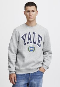 Sudadera gris con "Yale" en letras mayúsculas de color marino, adornada con un diseño de escudo. Mangas largas con puños acanalados y cuello redondo.
