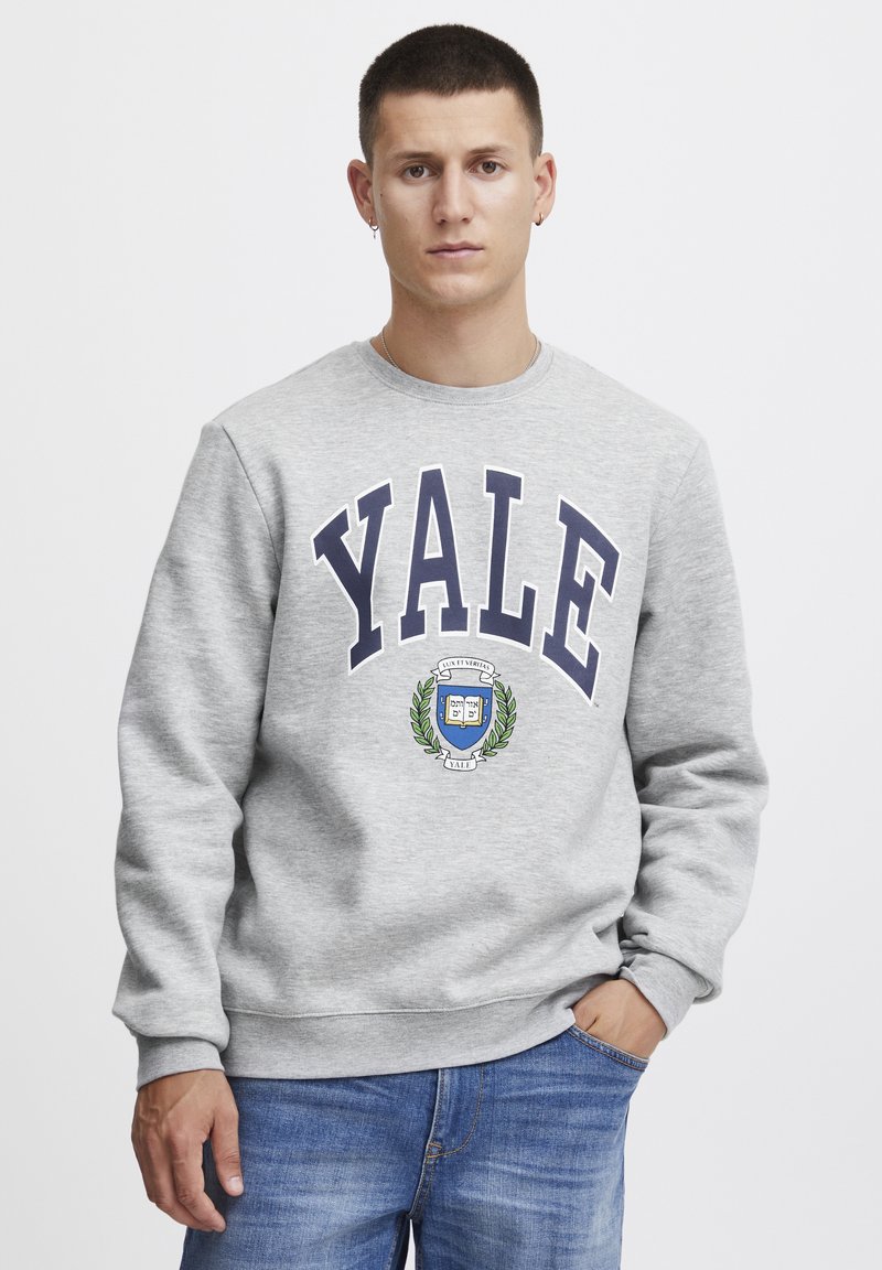 Sudadera gris con "Yale" en letras mayúsculas de color marino, adornada con un diseño de escudo. Mangas largas con puños acanalados y cuello redondo.