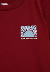 Bordo majica s kvadratnim printom i plavim dizajnom sunca te natpisom "BLUE RIDGE DENIM" u bijeloj boji. Izrađena od mekanog i glatkog materijala.