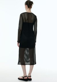Langes schwarzes Netzkleid mit runder Halslinie, langen Ärmeln und halbtransparenter Struktur, das unterhalb des Knies endet und mit einem detaillierten offenen Gewebe gefertigt ist.