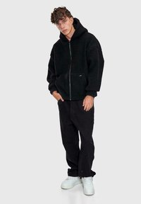 Giovane uomo che indossa una giacca in pile con cappuccio nera aperta con zip, pantaloni neri larghi e sneakers bianche, in piedi contro uno sfondo chiaro e semplice.