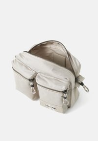 Light beige nylon bag with two front zip pockets, silver hardware, and a top zipper. Features a logo patch on the front. Soft texture.