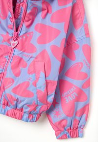 Veste imperméable rose et bleue avec des motifs de cœurs et d'étoiles, des poignets élastiques et une fermeture éclair à l'avant avec un tirette en forme de cœur.