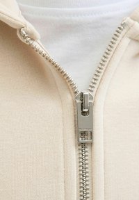 Zbliżenie na beżowy suwak z metalowym uchwytem w kolorze srebrnym, wyoblikowanym napisem "JACK & JONES" na jasnym ubraniu.