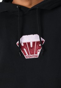 Sort hættetrøje med et diamantformet, broderet logo. Logoet har "HUF" i rødt og "OUTERNATIONAL" nedenunder i hvidt.
