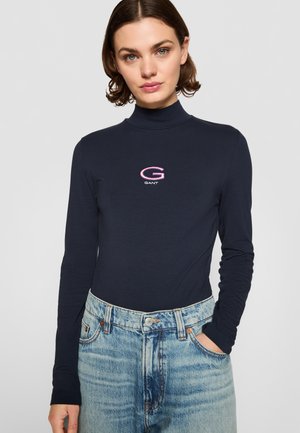 Marineblauw langetermijn coltrui met een geborduurd "GANT" logo in roze en wit op de borst, gecombineerd met hoogwrigte lichtblauwe jeans.