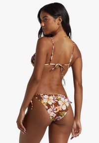 Bruine bloemenbikini met verstelbare bandjes, met grote witte en pastelbloemen, gestrikte zijkanten en een lage taille. Gladde textuur.