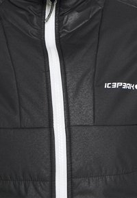 Svart isolerad jacka med en vit dragkedja, quiltad yta och logotyp på bröstet. Materialet verkar mjukt och lätt.