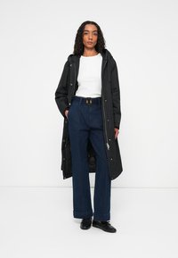 Zwarte lange jas met capuchon, witte lange mouw top en blauwe wijde denim broek. De broek heeft een hoge taille en opgerolde manchetten. Zwarte schoenen.