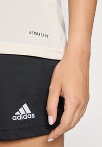 Lys beige mesh sports top med "AEROREADY" label, parret med sorte shorts med hvid Adidas-logo; glat tekstur, tætsiddende design.