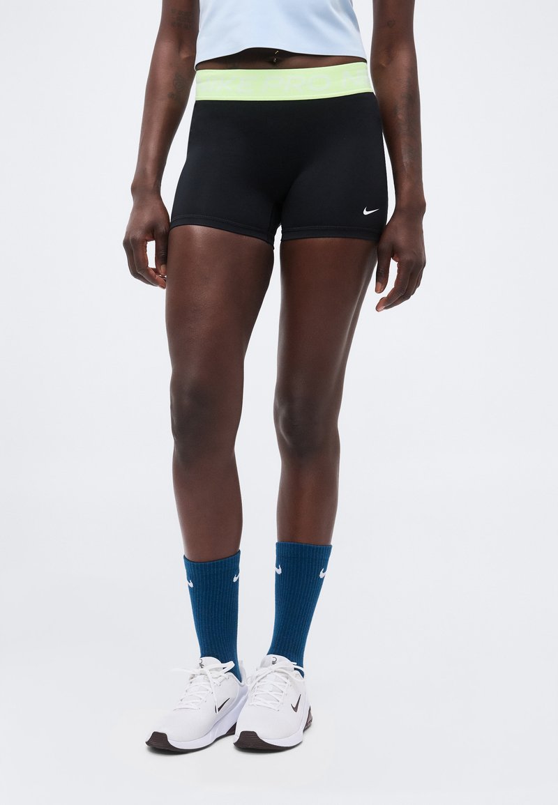 Άτομο που φοράει μαύρα σορτς Nike Pro, μπλε κάλτσες Nike και λευκά παπούτσια τρεξίματος Nike σε απλό φόντο.