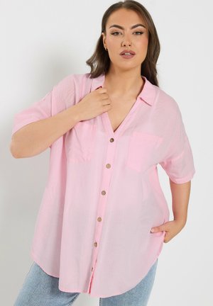 Frau trägt lockeres, hellrosa Hemd mit kurzen Ärmeln und blauen Jeans, posiert mit einer Hand auf der Brust und der anderen in der Hosentasche.