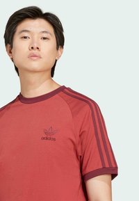 Crvena pamučna majica s okruglim izrezom, bordo akcentima i tri bordo pruge na svakoj rukavu. Sadrži izvezeni Adidas logo na grudima.