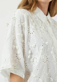 Camicia bianca di cotone con ricami floreali, motivi ad occhiello, colletto e maniche corte e ampie. Dettagli testurizzati in tutto il capo.