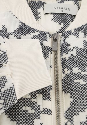 Witte jas met zwart kruissteekpatroon, zilveren rits met 'NK' gravure, geribbelde kraag en manchetten, en 'NUKUS S' label aan de binnenkant van de kraag.