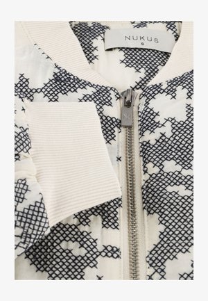 Witte jas met zwart kruissteekpatroon, zilveren rits met 'NK' gravure, geribbelde kraag en manchetten, en 'NUKUS S' label aan de binnenkant van de kraag.