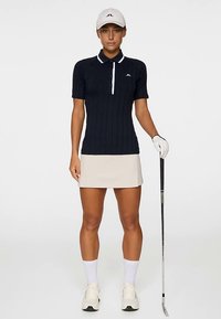 Granatowa koszulka polo z prążkowaną fakturą, białym kołnierzykiem i guzikami, zestawiona z jasnobeżową spódnicą. W ręku kij golfowy, na głowie biała czapka i rękawiczka.