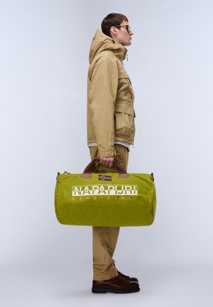 Man in een zandkleurig jasje en broek die een grote groene Napapijri reistas vasthoudt, zijwaarts staand tegen een effen lichte achtergrond.