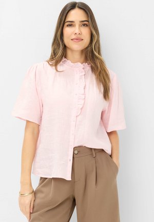 Frau trägt eine hellrosa Bluse mit Rüschen und Knopfleiste sowie eine hochtaillierte beige Hose, steht mit einer Hand in der Tasche vor einem schlichten Hintergrund.